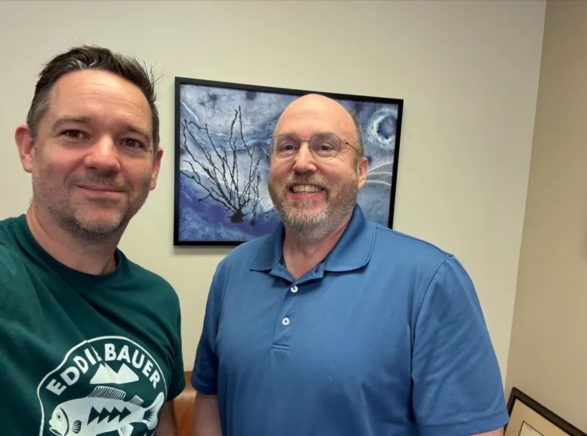 Sean Bendall (left), wearing a green T-shirt with a fish and “Eddie Bauer” in white, and Thomas Montine (right), in a blue polo shirt, poses in front of a blue painting and neutral-colored walls.