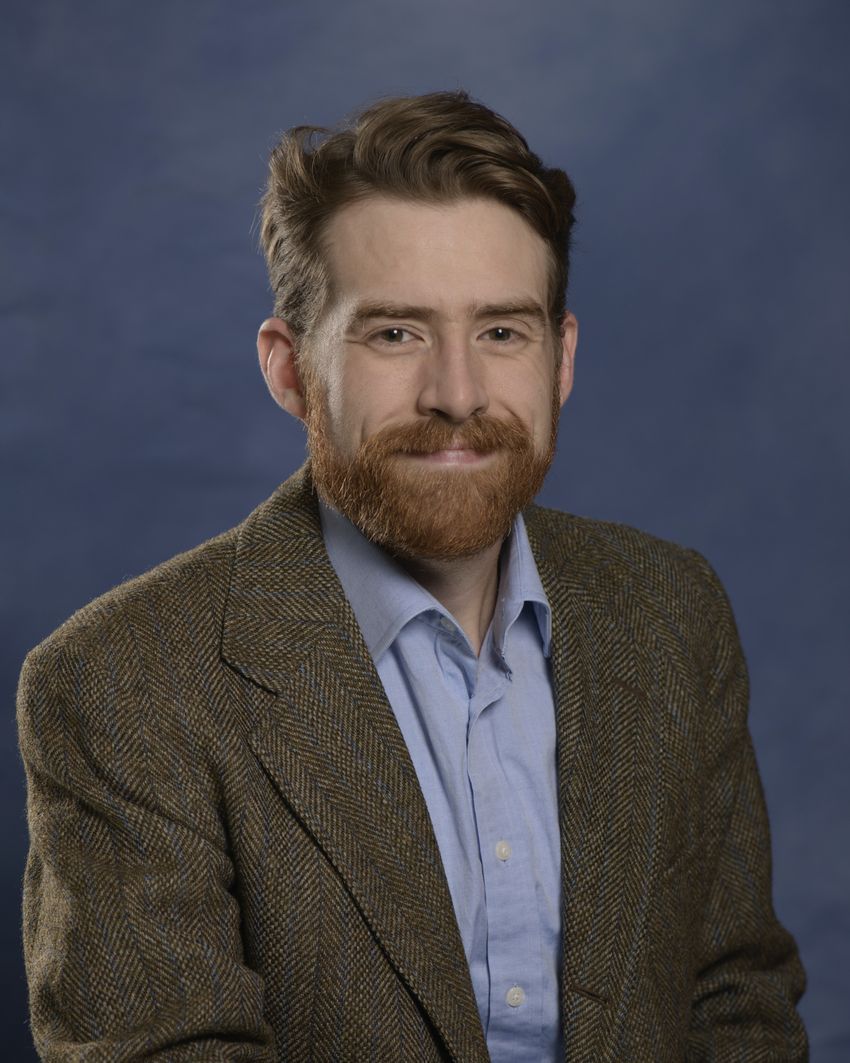 Adam Dolezal poses in front of a dark blue background. He’s wearing a light blue shirt and a dark brown patterned suit.