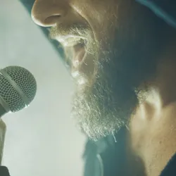 Photograph of the bottom half of a man’s face while he sings into a microphone. 