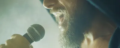 Photograph of the bottom half of a man’s face while he sings into a microphone. Photograph of the bottom half of a man’s face while he sings into a microphone.