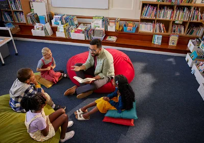 Image of a male teacher reading a book to a group of children while sitting on the floor of a school library.