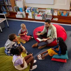 Image of a male teacher reading a book to a group of children while sitting on the floor of a school library.