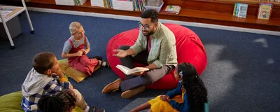 Image of a male teacher reading a book to a group of children while sitting on the floor of a school library. Image of a male teacher reading a book to a group of children while sitting on the floor of a school library.