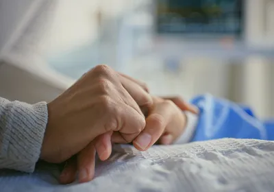 An individual with cancer holding hands with a bedside caregiver. An individual with cancer holding hands with a bedside caregiver.
