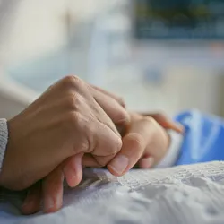 An individual with cancer holding hands with a bedside caregiver.