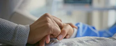 An individual with cancer holding hands with a bedside caregiver. An individual with cancer holding hands with a bedside caregiver.