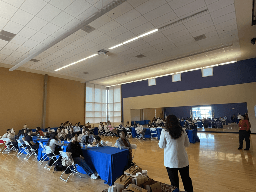 Young adults sitting at tables arranged in a U shape with blue table cloths listen to graduate students standing at the front of the room to hear about graduate school as part of a science outreach event, Café con Pan Dulce. Young adults sitting at tables arranged in a U shape with blue table cloths listen to graduate students standing at the front of the room to hear about graduate school as part of a science outreach event, Café con Pan Dulce.