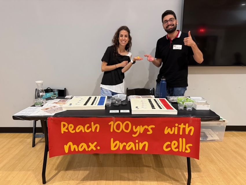 Photograph of two people, a woman with medium-length curly hair and a man with short dark hair, standing behind a table with microscope slides of brain slices. A red banner reads “Reach 100yrs with max. brain cells”. This table was at a science outreach event, the Amazing Brain Carnival, hosted the St. Louis Neuroscience Outreach group. Photograph of two people, a woman with medium-length curly hair and a man with short dark hair, standing behind a table with microscope slides of brain slices. A red banner reads “Reach 100yrs with max. brain cells”. This table was at a science outreach event, the Amazing Brain Carnival, hosted the St. Louis Neuroscience Outreach group.