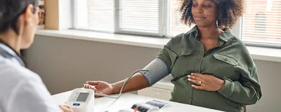 A pregnant Black woman with tightly coiled hair sits at a desk in a doctor’s office getting her blood pressure taken. A pregnant Black woman with tightly coiled hair sits at a desk in a doctor’s office getting her blood pressure taken.