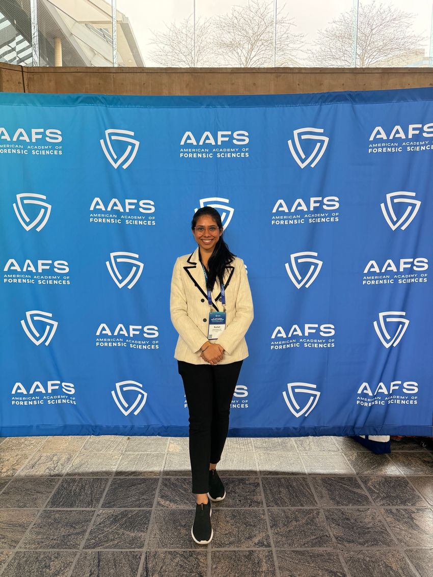 Rushali Dargan stands in front of a blue set with the “American Academy of Forensic Sciences” name and logo. She’s wearing a beige jacket, black pants, black sneakers, and glasses. Rushali Dargan stands in front of a blue set with the “American Academy of Forensic Sciences” name and logo. She’s wearing a beige jacket, black pants, black sneakers, and glasses.
