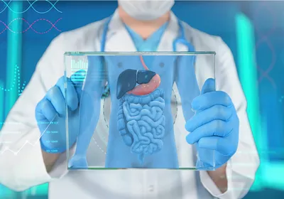 A doctor in a white coat, blue gloves, and surgical mask holds a clear screen showing a gastrointestinal tract and some medical data. 
