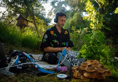 Image of Tarun Nayar sitting on the ground with a modular synthesizer in his lab. There are mushrooms in the foreground.