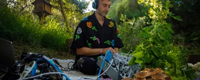 Image of Tarun Nayar sitting on the ground with a modular synthesizer in his lab. There are mushrooms in the foreground. Image of Tarun Nayar sitting on the ground with a modular synthesizer in his lab. There are mushrooms in the foreground.