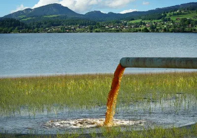 Brightly colored water, representing chemical waste contamination by PFAS, drains into a natural body of water. 