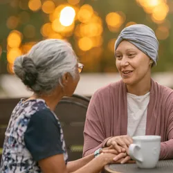 A young female cancer patient spending time with a friend. They are sitting outside, enjoying tea and conversation, as the sun sets behind them.
