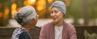 A young female cancer patient spending time with a friend. They are sitting outside, enjoying tea and conversation, as the sun sets behind them.