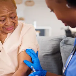  An older woman gets a vaccine in her arm from a doctor wearing blue scrubs and blue gloves.