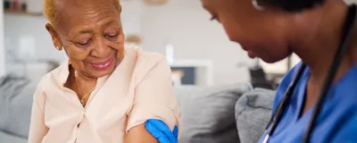 An older woman gets a vaccine in her arm from a doctor wearing blue scrubs and blue gloves. An older woman gets a vaccine in her arm from a doctor wearing blue scrubs and blue gloves.