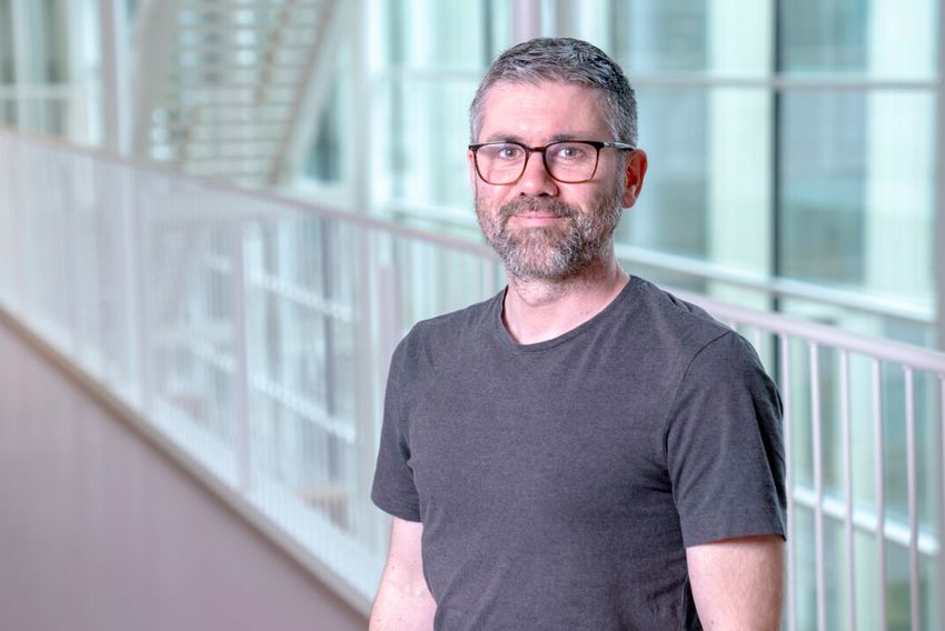 René Aquarius, wearing a dark grey heathered tee and glasses, poses inside a building with stairs and glass windows. René Aquarius, wearing a dark grey heathered tee and glasses, poses inside a building with stairs and glass windows.