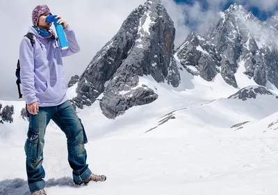 A mountain climber stands in front of snowy mountains and holds a blue canister of portable supplemental oxygen to his face to relieve symptoms of hypoxia due to the high altitude. A mountain climber stands in front of snowy mountains and holds a blue canister of portable supplemental oxygen to his face to relieve symptoms of hypoxia due to the high altitude.
