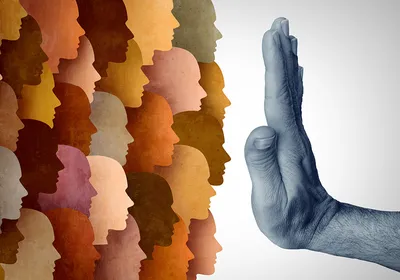 Representative image of travel bans. One the left are silhouettes of human faces in colors ranging from cream to tan to brown. A hand extending from the right is held up in a “stop” signal in front of them in the middle of the image. 