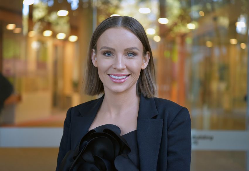 Joy Wolfram, an extracellular vesicle researcher, is wearing a black shirt and jacket and smiling in front of a background filled with yellow and orange lights. Joy Wolfram, an extracellular vesicle researcher, is wearing a black shirt and jacket and smiling in front of a background filled with yellow and orange lights.