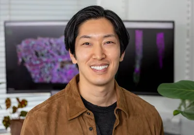 Image of Tatsuya Nobori. He smiles at the camera and wears a black shirt underneath a brown jacket. There are computer monitors in the background that depict colorful images. 