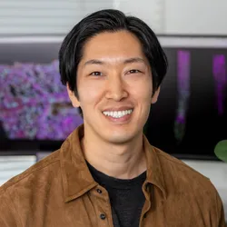 Image of Tatsuya Nobori. He smiles at the camera and wears a black shirt underneath a brown jacket. There are computer monitors in the background that depict colorful images. 