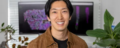 Image of Tatsuya Nobori. He smiles at the camera and wears a black shirt underneath a brown jacket. There are computer monitors in the background that depict colorful images. Image of Tatsuya Nobori. He smiles at the camera and wears a black shirt underneath a brown jacket. There are computer monitors in the background that depict colorful images.