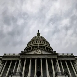 The US capitol building is shown from below at an angle, with a cloudy sky in the background.