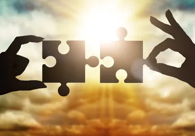 Hands collect gear in a puzzle against the sky in the sunset, representing academia-industry partnership.