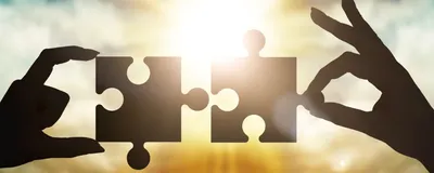 Hands collect gear in a puzzle against the sky in the sunset, representing academia-industry partnership.