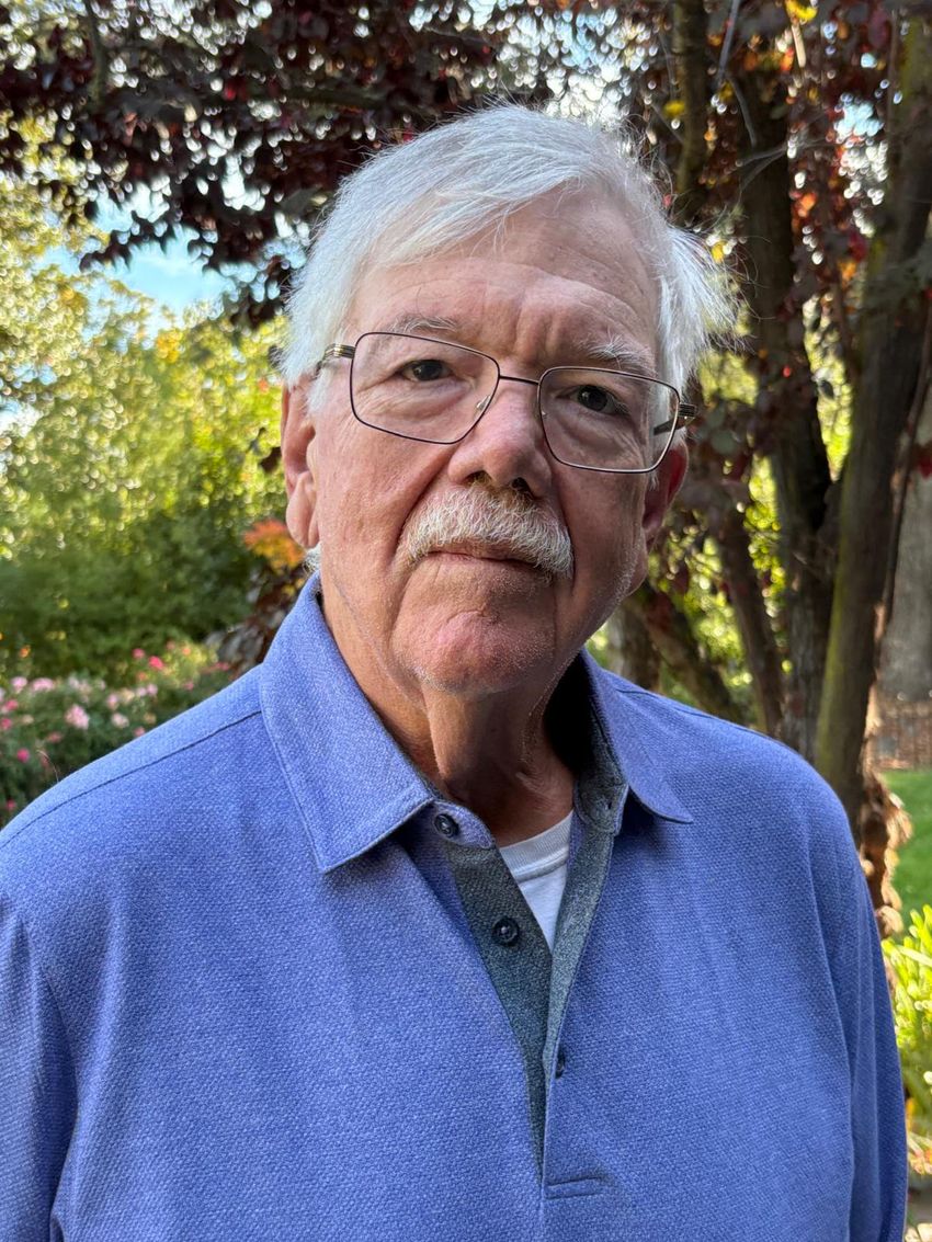 Henry (Hank) Greely, wearing a light blue collared shirt and glasses, stands outdoors in front of trees.