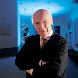 James Watson, wearing a black jacket over a shirt and tie, poses in a room with a model of the DNA double helix, which he co-discovered in 1953, in the background. 