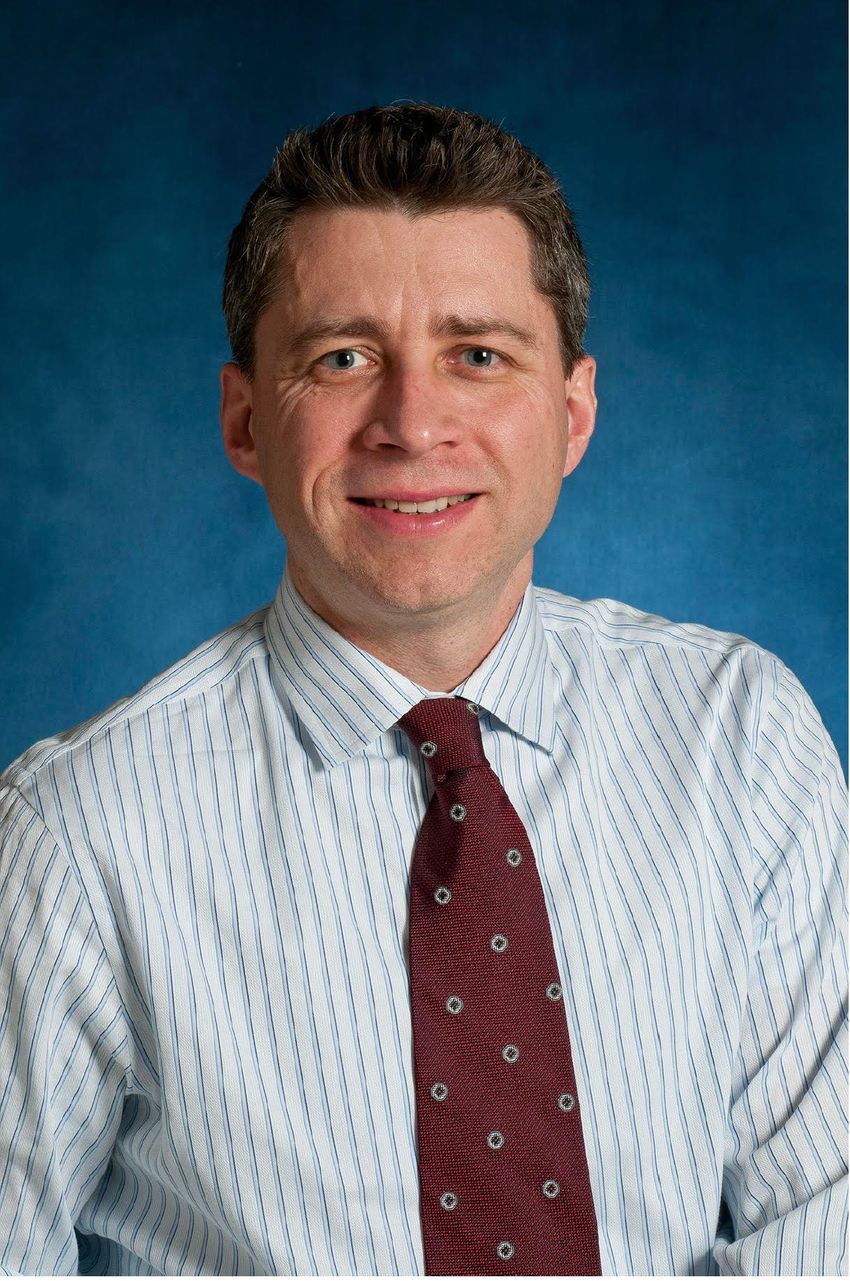Edward Harhaj smiles in front of a blue background, wearing a white shirt with blue stripes and a red tie with white circles. Edward Harhaj smiles in front of a blue background, wearing a white shirt with blue stripes and a red tie with white circles.