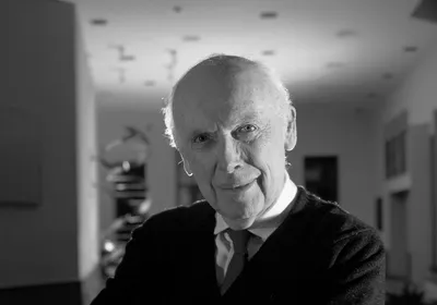 Black-and-white image of James Watson, wearing a black jacket over a shirt and tie, poses in a room with a model of the DNA double helix, which he co-discovered in 1953, in the background.