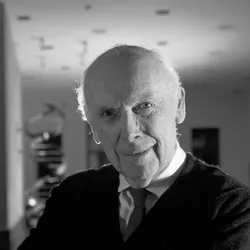 Black-and-white image of James Watson, wearing a black jacket over a shirt and tie, poses in a room with a model of the DNA double helix, which he co-discovered in 1953, in the background.