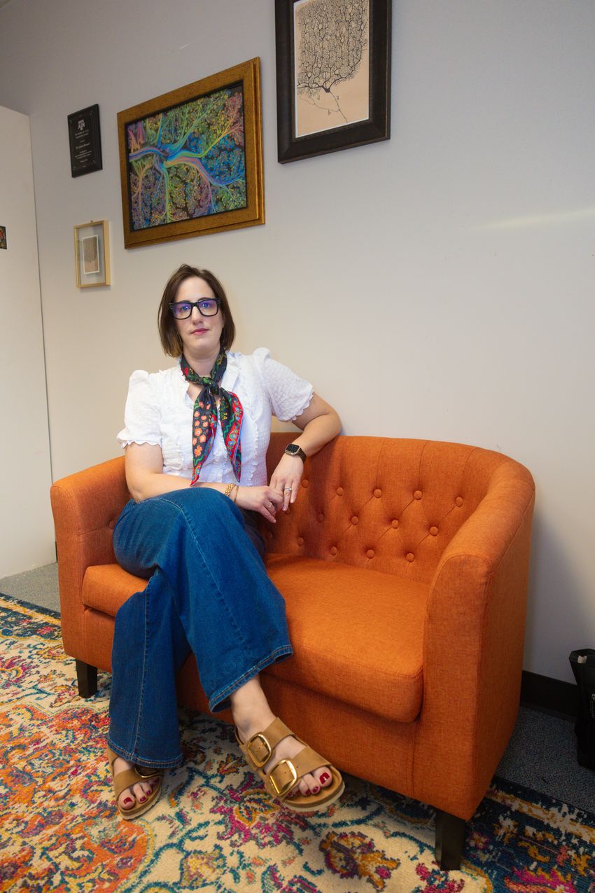 Jessica Bernard, a cognitive neuroscientist, wears a white top and jeans while sitting on an orange couch with neuroscience-inspired art on the wall behind her.