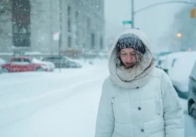 A woman walks under snowfall on a city street. Signifies that some people feel cold temperatures more than others.