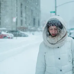 A woman walks under snowfall on a city street. Signifies that some people feel cold temperatures more than others.