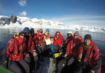 Nine people wearing red coats sit in a small inflatable boat on the ocean in front of an Antarctic Fjord. These are guests joining in the citizen science project FjordPhyto to collect water samples to study phytoplankton.