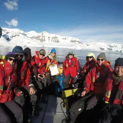 Nine people wearing red coats sit in a small inflatable boat on the ocean in front of an Antarctic Fjord. These are guests joining in the citizen science project FjordPhyto to collect water samples to study phytoplankton.