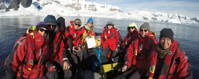 Nine people wearing red coats sit in a small inflatable boat on the ocean in front of an Antarctic Fjord. These are guests joining in the citizen science project FjordPhyto to collect water samples to study phytoplankton.