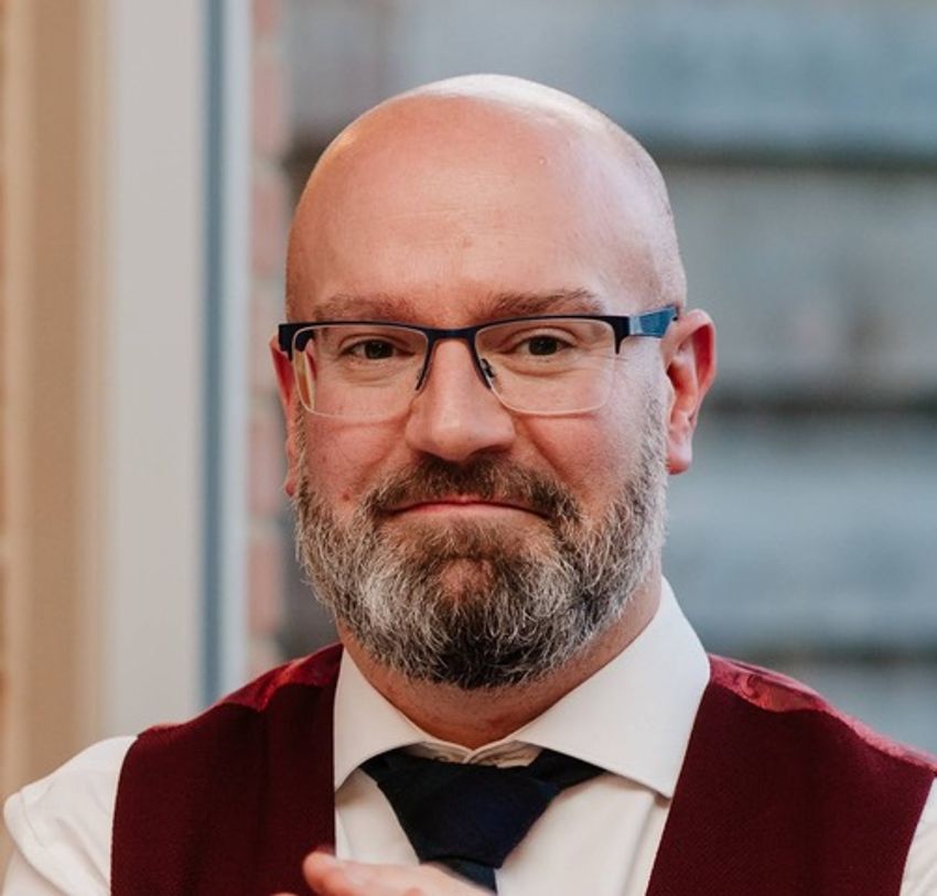 Oliver Smith, wearing a white shirt, black tie, red vest, and glasses, poses in front of a blurred background. Oliver Smith, wearing a white shirt, black tie, red vest, and glasses, poses in front of a blurred background.
