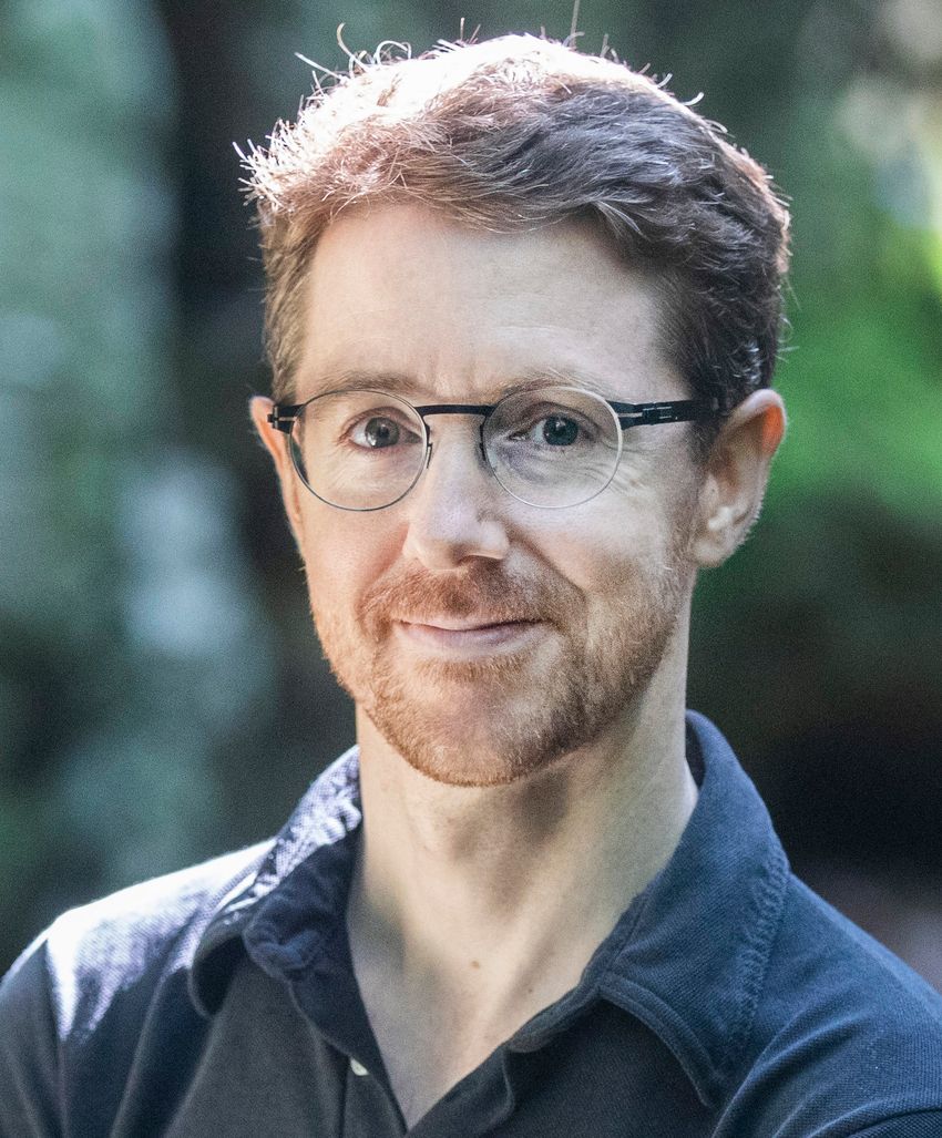 Martin Picard, wearing a dark collared shirt and glasses, poses in front of a blurred background.