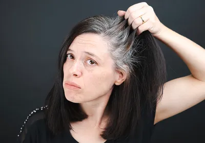 A woman pulls on her dark hair, showing graying roots. She looks frustrated, representing how stress can cause hair to go gray prematurely. 