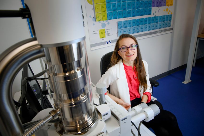 Maria McNamara poses in a laboratory. She’s wearing a pink shirt, white jacket, black pants, and glasses.