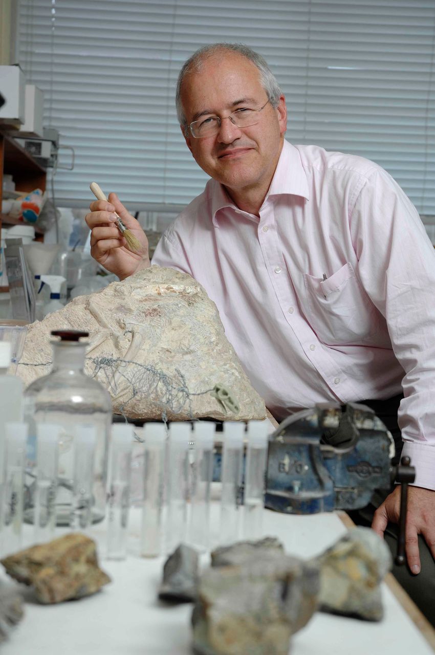Mike Benton sits by his lab bench, holding a brush. On the table next to him, there are several rocks, test tubes, and a small skeletal model of a dinosaur.