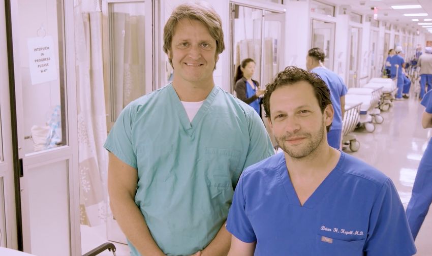 Alexandar Charney, a psychiatrist, stands in green scrubs to the left of Brian Kopell, a neurosurgeon, who is wearing blue scrubs. Charney has short light hair and Kopell has short dark hair. They are standing in a hospital hallway with other personnel in scrubs in the background.