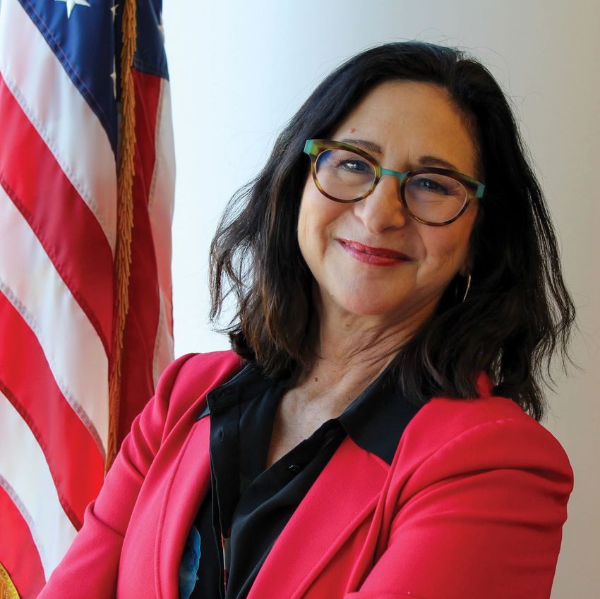 Photograph of Dorit Donoviel, the executive director of the Translational Research Institute for Space Health at the Baylor College of Medicine. Donoviel has medium-length dark hair and is wearing a salmon blazer in front of a white wall with an American flag on the left of the image. She is smiling at the camera.